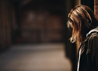 Image: A woman stands with her back to a wall, looking down at the ground