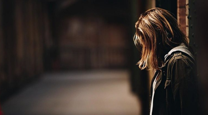 Image: A woman stands with her back to a wall, looking down at the ground