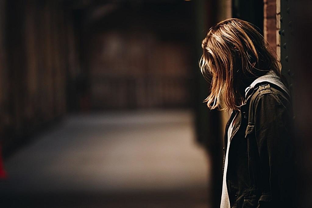 Image: A woman standing with her back to a wall, looking down at the ground