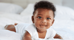Image description: a baby smiling and wearing a white onesie while doing tummy time on a bed. (Tummy Time Without Tears: Tips From a Pediatric Physical Therapist Brittany Aquart Contributor Miami Mom Collective)