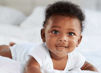 Image description: a baby smiling and wearing a white onesie while doing tummy time on a bed. (Tummy Time Without Tears: Tips From a Pediatric Physical Therapist Brittany Aquart Contributor Miami Mom Collective)
