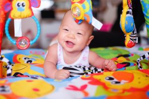 Image Description: a baby is smiling while doing tummy time on a brightly colored mat. Colorful toys are hanging from a play gym in front of the baby. (Tummy Time Without Tears: Tips From a Pediatric Physical Therapist Brittany Aquart Contributor Miami Mom Collective)