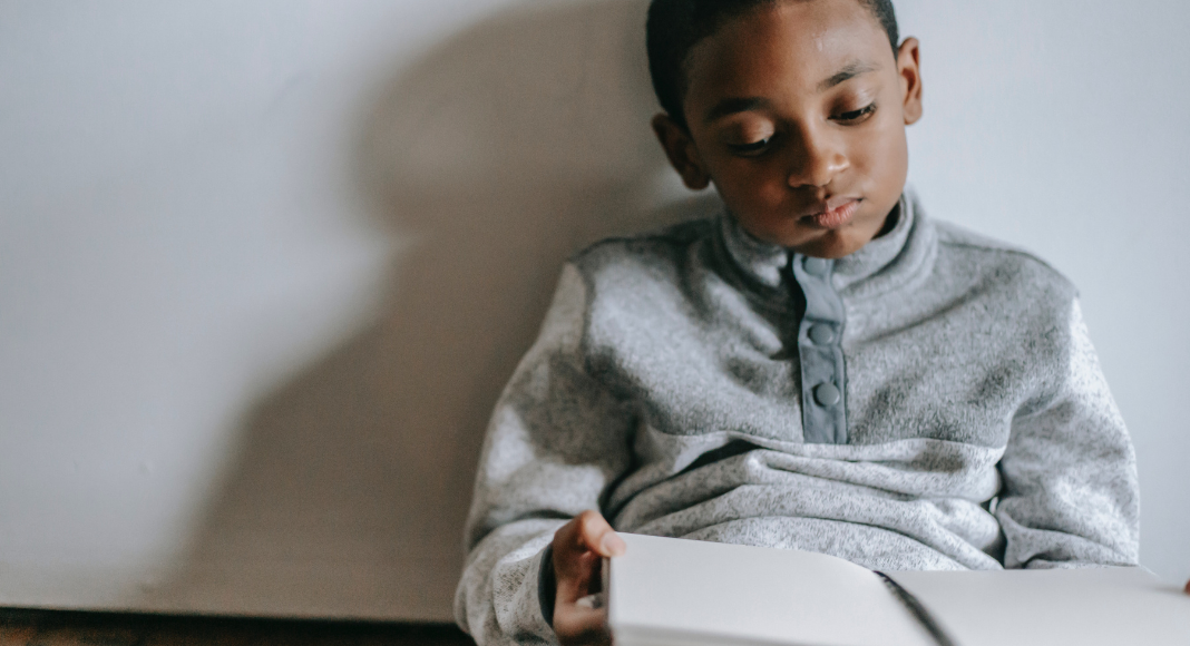 Image: A boy sitting with a book in his lap (Stuck Inside? Try These Gross Motor Games! Brittany Aquart Contributor Miami Mom Collective)