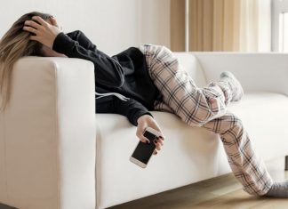 A burned out woman lies on the couch, holding her head in one hand and her phone in the other