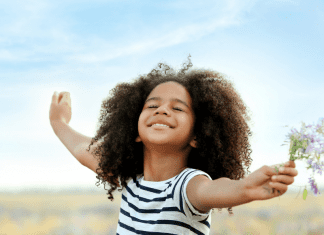 A young girl stands in a field with her arms spread wide, eyes closed and smiling. She is holding a bunch of wildflowers and the wind is blowing her hair.