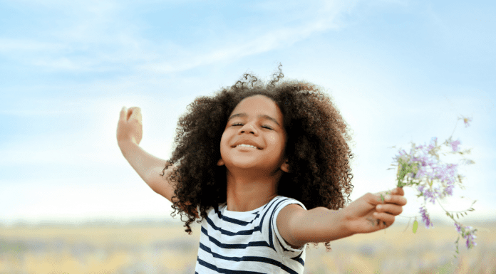 A young girl stands in a field with her arms spread wide, eyes closed and smiling. She is holding a bunch of wildflowers and the wind is blowing her hair.