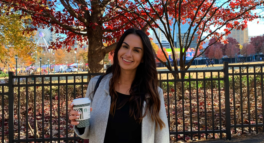 Jessica, holding a cup of coffee at a park with fall leaves behind her