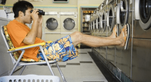 A young man sits in a chair with a pair of binoculars, watching his laundry at the laundromat