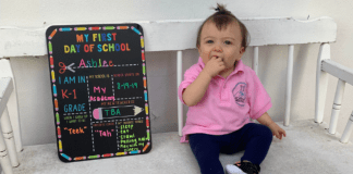 A toddler sits next to a My First Day of School board