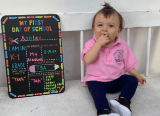 A toddler sits next to a My First Day of School board