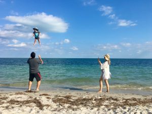 Image: A woman takes a photo of a dad playfully throwing and catching his toddler at the beach