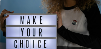 Image: A woman with an "I voted" sticker holds a letterboard that reads, "Make Your Choice"