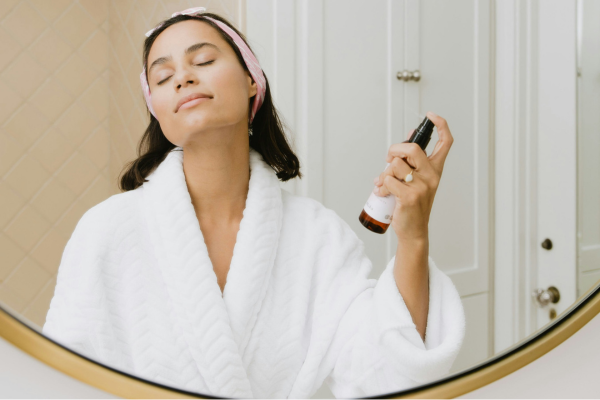 Image: A woman stands in front of a mirror and applies a skincare spray product