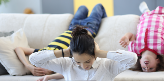 Image: A mom sits in front of a laptop with her hands on her ears, while two children hang upside down on the couch