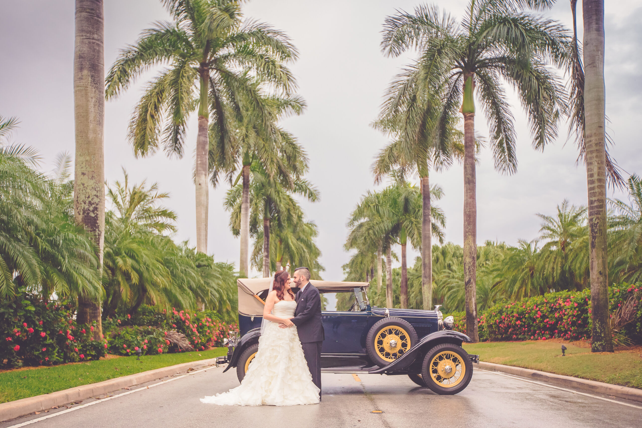 Bride is dressed in a light cream wedding dressing being held by her husband to be in a black suit and black leather sneakers. They are leaning on a navy blue 1930s Model T car. They are surrounded by towering palm trees in Miami, FL. 