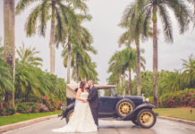 Bride is dressed in a light cream wedding dressing being held by her husband to be in a black suit and black leather sneakers. They are leaning on a navy blue 1930s Model T car. They are surrounded by towering palm trees in Miami, FL.